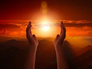 An image of two hands reaching up to the sky with the sun in-between them to reference how problems with spirituality can affect a person's health, or make it harder for people to heal.