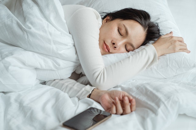 Why sleep causes junk food cravings with a picture of a woman sleeping in white sheets with white pajamas.